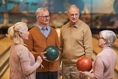 Waist up portrait of group of senior people holding bowling balls and chatting while enjoying active entertainment at bowling alley, copy space