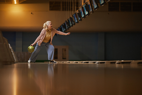 Wide angle side view at senior woman playing bowling alone while enjoying active entertainment at bowling alley, copy space