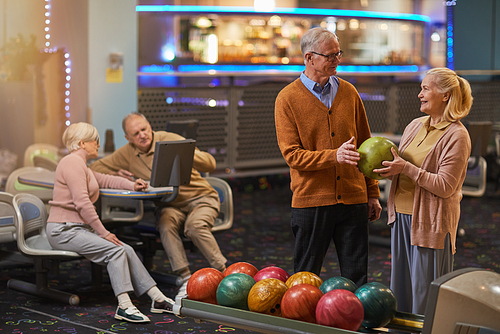 Portrait of smiling senior couple playing bowling together with friends in background while enjoying active entertainment at bowling alley, copy space