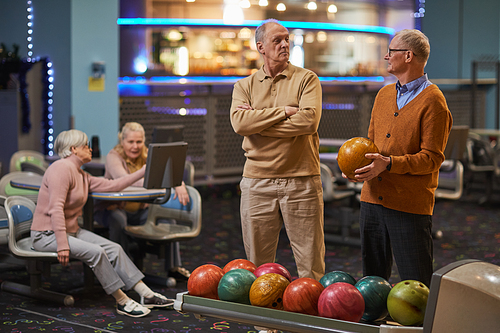 Portrait of two senior men playing bowling together with friends in background while enjoying active entertainment at bowling alley, copy space