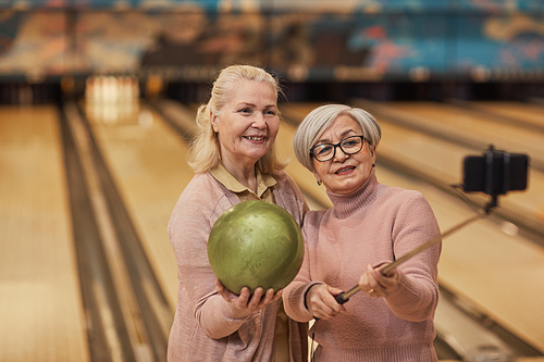 Waist up portrait of two senior women taking selfie photos while playing bowling and enjoying active entertainment at bowling alley, copy space