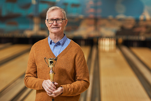 Waist up portrait of smiling senior man holding trophy and looking at camera while standing in bowling alley after winning match, copy space