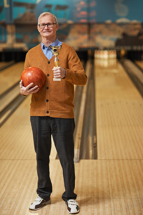 Vertical full length portrait of smiling senior man holding trophy and bowling ball while posing at bowling alley after winning match, copy space