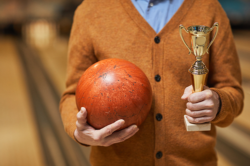 Cropped shot of unrecognizable mature man holding trophy and bowling ball while posing at bowling alley after winning match, copy space