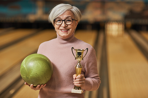 Waist up portrait of smiling senior woman holding trophy and looking at camera while standing in bowling alley after winning match, copy space