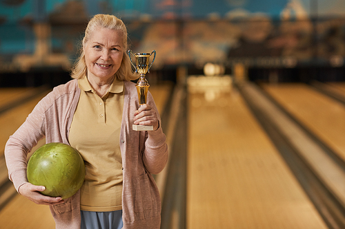 Waist up portrait of smiling mature woman holding trophy and looking at camera while standing in bowling alley after winning match, copy space