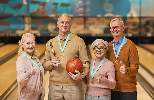 Waist up portrait of smiling senior team wearing medals holding trophy and looking at camera while standing in bowling alley after winning match