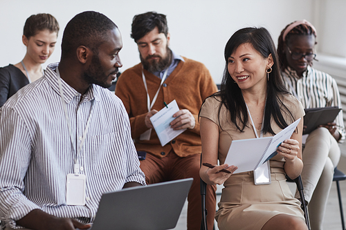 Multi-ethnic group of people in audience at business conference focus on smiling man and woman talking while sitting on chairs in row