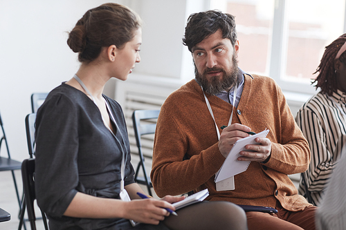 Group of people in audience at business conference focus on bearded man talking to woman while sitting on chairs