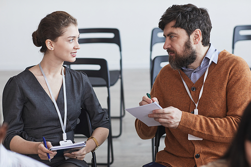 Front view portrait of two people in audience at business conference, man and woman talking and taking notes while sitting on chairs in row