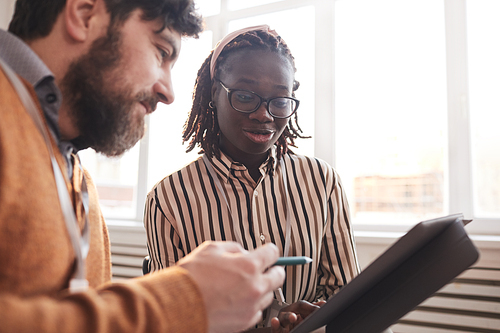 Portrait of young African-American woman talking to colleague and looking at tablet while discussing business project in office