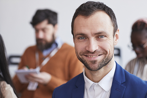 Head and shoulders portrait of smiling businessman looking at camera with audience at business conference in background, copy space