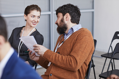 Portrait of bearded businessman talking to smiling young woman while sitting in audience at business conference, copy space