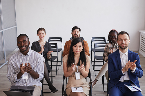 High angle view at multi-ethnic group of people looking at camera and applauding while sitting in audience at business conference or seminar, copy space