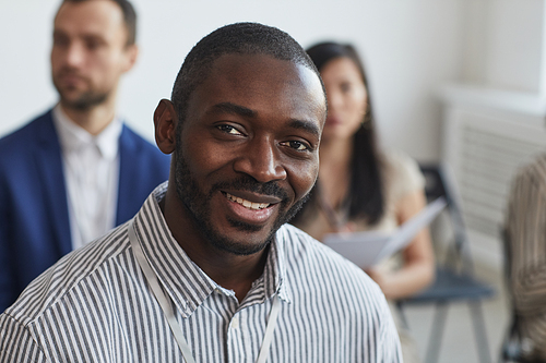 Head and shoulders portrait of African-American businessman looking at camera and smiling while sitting in audience at business conference or seminar, copy space