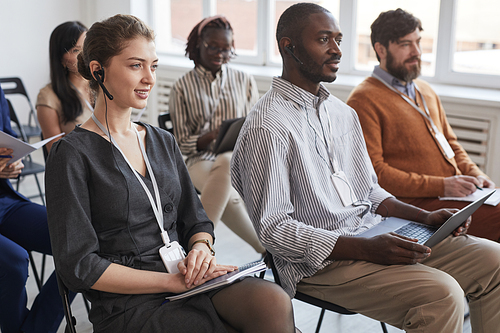 Multi-ethnic group of people sitting on chairs in audience at business conference or seminar, focus on young woman wearing headset in foreground