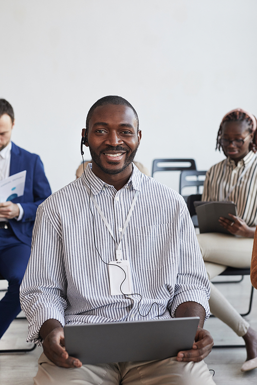 Vertical portrait of smiling African-American businessman wearing headset and holding laptop while sitting in audience at business conference or seminar, copy space