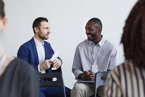 Portrait of two smiling businessmen talking while sitting on chairs in audience at business conference or seminar, copy space