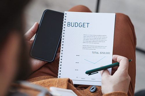 High angle close up of unrecognizable man holding note pad with BUDGET planner and smartphone, copy space