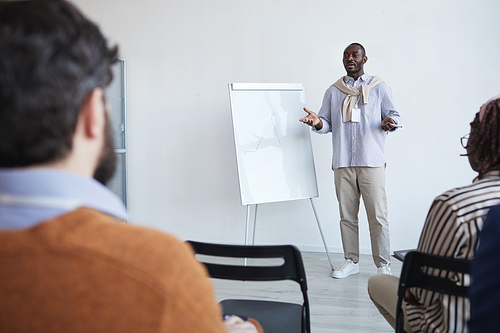 Wide angle portrait of African-American business coach talking to audience at conference or education seminar while standing by whiteboard and gesturing, copy space