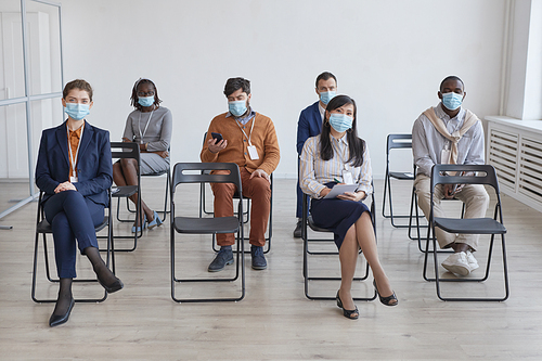 Full length multi-ethnic group of business people wearing masks and social distancing while sitting on chairs in audience at business conference or seminar