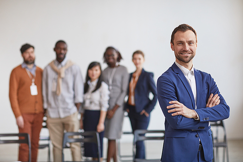 Waist up portrait of successful businessman standing with arms crossed against multi-ethnic team in background, copy space