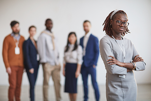 Waist up portrait of successful African-American businesswoman standing with arms crossed against multi-ethnic team in background, copy space