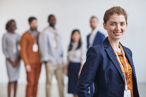 Waist up portrait of successful businesswoman smiling at camera while standing with multi-ethnic team in background, copy space