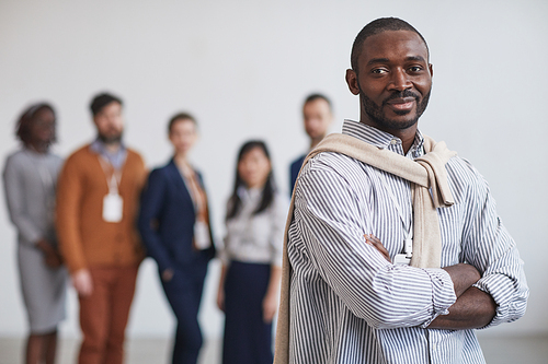 Waist up portrait of successful African-American businessman smiling at camera while standing with multi-ethnic team in background, copy space