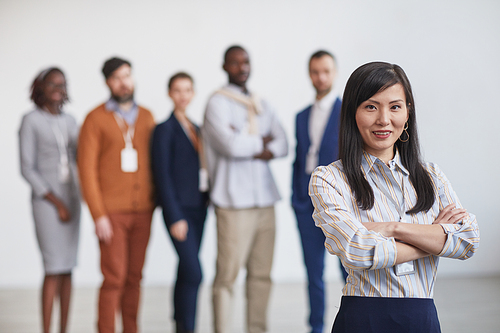 Waist up portrait of successful Asian businesswoman smiling at camera while standing with multi-ethnic team in background, copy space