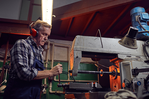 Serious senior operator wearing headphones standing at the lathe and operating it in the factory