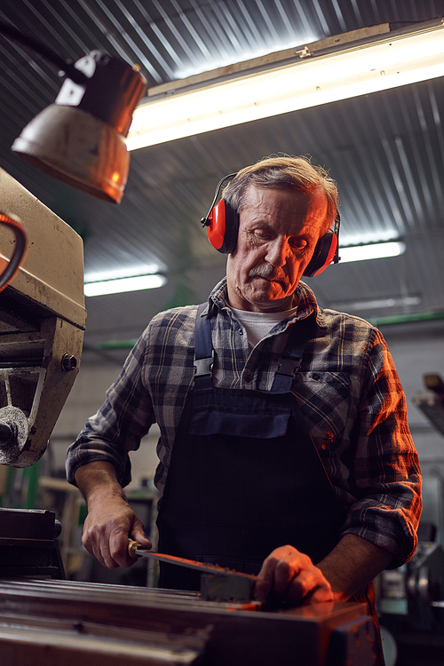 Senior manual worker in headphones standing near the machine and sawing the metal with saw