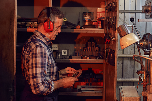 Senior worker wearing overalls and headphones choosing a disk for grinder while standing in the workshop