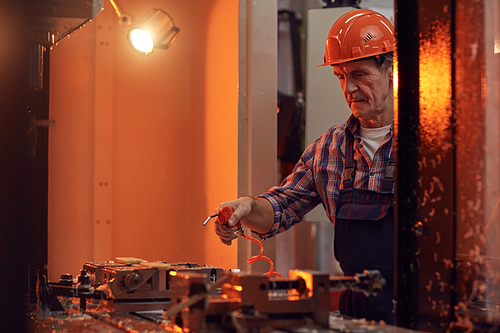 Mature engineer in work helmet using special equipment with wires while working at the lathe