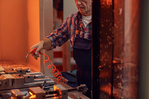Close-up of mature worker in work wear holding work tool and blowing on metal details while working at the lathe