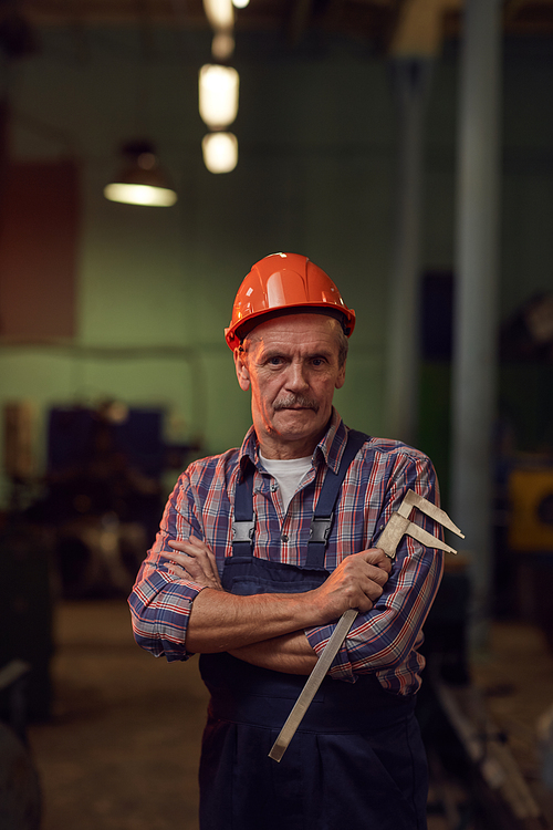 Portrait of senior manual worker in work helmet holding measuring equipment and looking at camera while standing in the factory