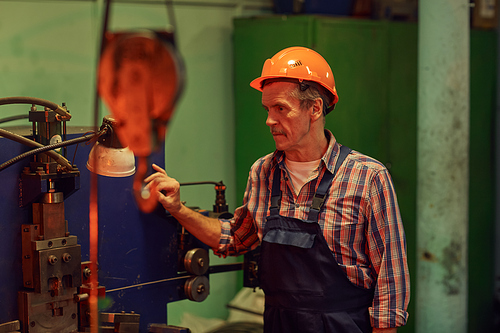 Senior mature worker in overalls and in work helmet standing at the lathe and watching for the process
