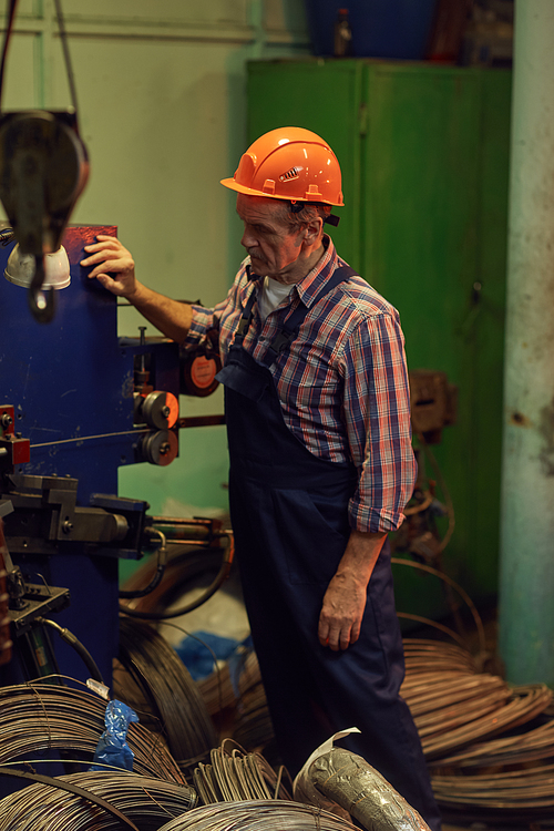 Mature engineer in work helmet standing at the lathe among cables and watching for the work of lathe