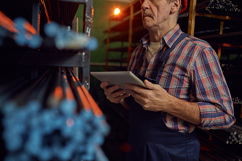 Close-up of mature foreman holding digital tablet and checking availability of metal pipes in warehouse