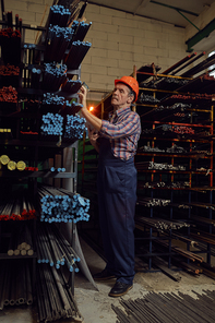 Senior worker in work wear and in helmet standing and examining the metal pipes on the shelves in the metal factory