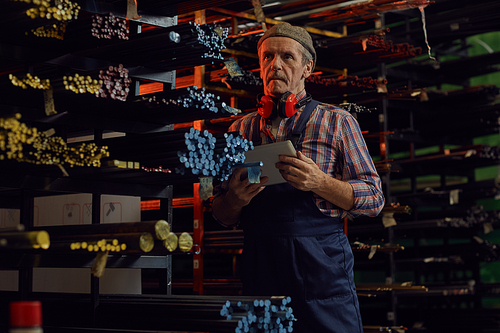 Mature manual worker in overalls using digital tablet and looking at colorful metal pipes on shelves