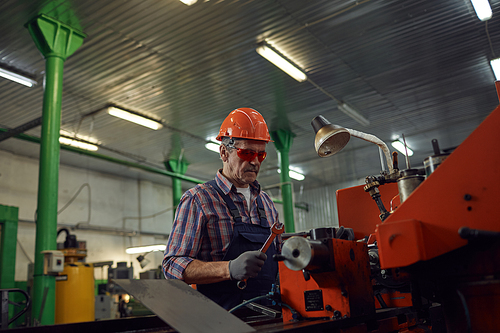 Mature mechanic in protective eyeglasses and in work helmet repairing the lathe with work tool