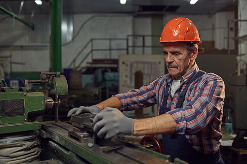Mature manual worker in work helmet and in gloves cutting the metal details with lathe in the factory