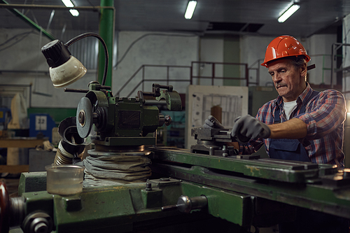 Serious mature engineer in work helmet standing at the lathe and cutting metal details in the metal factory