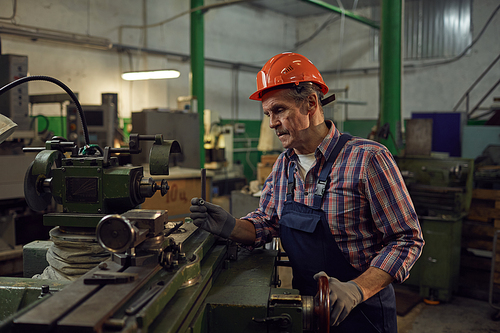 Mature man in overalls and in work helmet using lathe to do his work in the plant