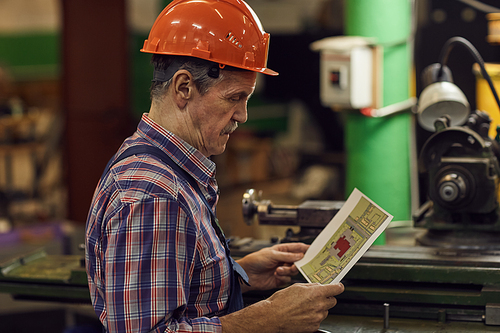 Mature manual worker in work helmet looking at plan in his hands before his work on lathe