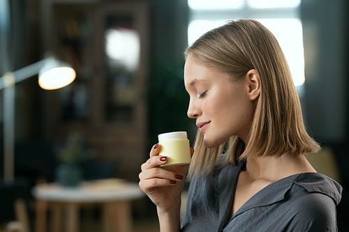 Young pretty female with blond hair enjoying smell of natural handmade cosmetic product in small plastic jar before applying it on her skin