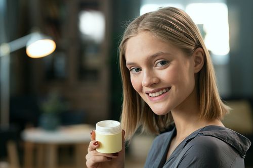 Young pretty female with blond hair holding small jar with natural handmade cosmetic product and looking at you with toothy smile