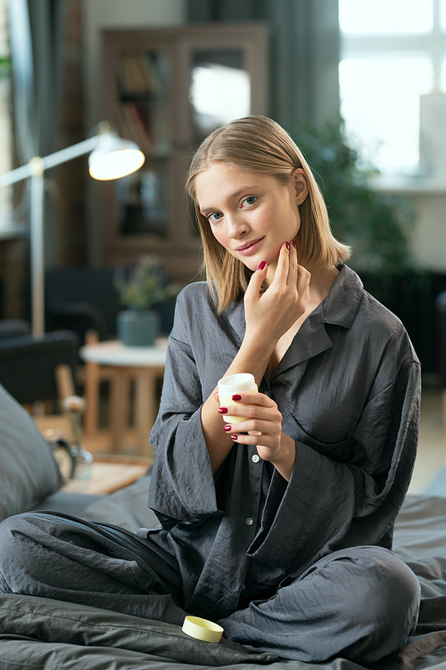 Young blond pretty female in grey pajamas applying natural handmade cosmetic product on her face while sitting on bed in the morning