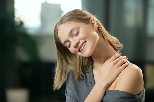 Young blond woman with toothy smile applying natural handmade cosmetic product on her neck and body and enjoying it in the morning